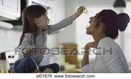 Stock Photo - Profile of playful daughter feeding mother spaghetti pasta in kitchen. Fotosearch