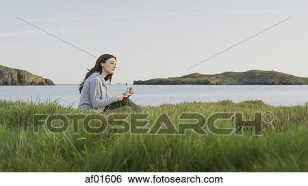 Stock Photograph - Profile of woman sitting in grassy field near lake in remote landscape under blue sky. Fotosearch