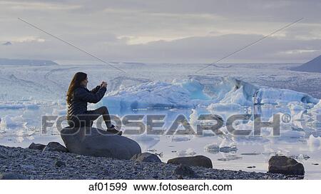 Stock Photo - Profile view of woman photographing remote landscape and frozen glaciers in still ocean under cloudy sky. Fotosearch