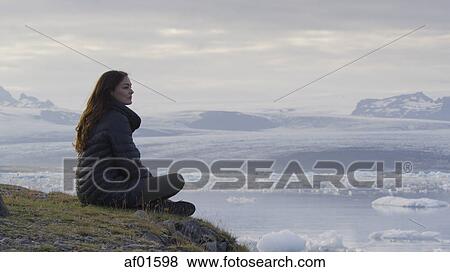 Profile view of woman sitting on grassy hilltop overlooking landscape and frozen glaciers in remote still ocean under cloudy sky View Large Photo Image Stock Photo - Profile view of woman sitting on grassy hilltop overlooking landscape and frozen glaciers in remote still ocean under cloudy sky. Fotosearch