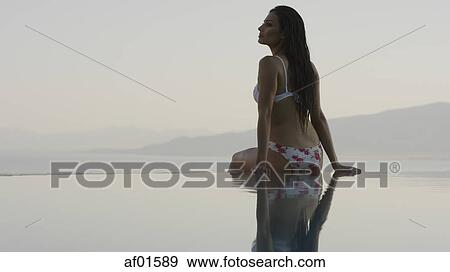 Rear view of woman in bikini sitting at edge of infinity pool overlooking horizon and remote landscape View Large Photo Image Stock Photo - Rear view of woman in bikini sitting at edge of infinity pool overlooking horizon and remote landscape. Fotosearch