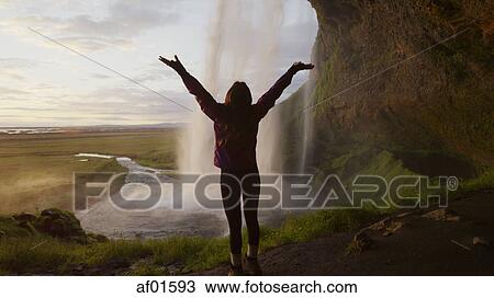 Rear view silhouette of woman with outstretched arms standing behind waterfall overlooking remote landscape View Large Photo Image Stock Image - Rear view silhouette of woman with outstretched arms standing behind waterfall overlooking remote landscape. Fotosearch