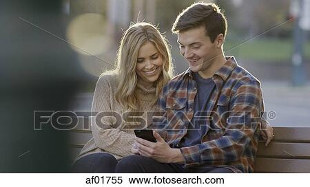 Selective focus view of smiling boyfriend and girlfriend sharing smartphone and sitting on bench View Large Photo Image Stock Photography - Selective focus view of smiling boyfriend and girlfriend sharing smartphone and sitting on bench. Fotosearch