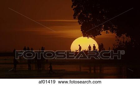 Silhouette of people standing on scenic beach under glowing sun in sunset sky View Large Photo Image Stock Photo - Silhouette of people standing on scenic beach under glowing sun in sunset sky. Fotosearch