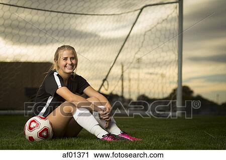 Stock Image - Smiling athlete sitting with ball in soccer goal under sunset sky. Fotosearch