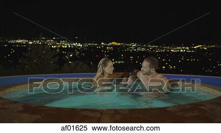 Stock Photography - Smiling couple sitting in illuminated hot tub overlooking cityscape at night. Fotosearch