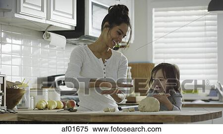 Stock Photography - Smiling mother teaching daughter to bake and roll dough in kitchen. Fotosearch