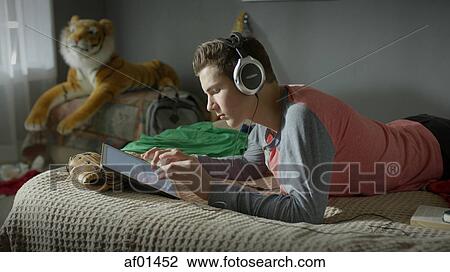 Teenage boy listening to headphones and using digital tablet on bed View Large Photo Image Stock Image - Teenage boy listening to headphones and using digital tablet on bed. Fotosearch