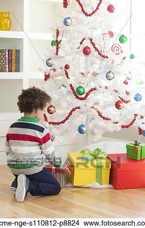 Picture - Young boy sitting under Christmas tree, picking up Christmas presents. Fotosearch