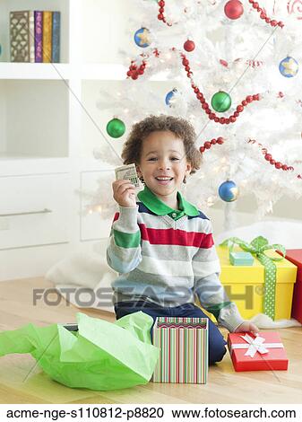 Stock Image - Young boy sitting under Christmas tree, holding dollar note. Fotosearch