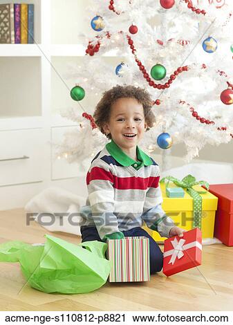 Stock Image - Young boy sitting under Christmas tree, opening Christmas presents. Fotosearch