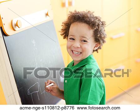 Stock Photograph - Young boy standing next to small blackboard, holding chalk. Fotosearch