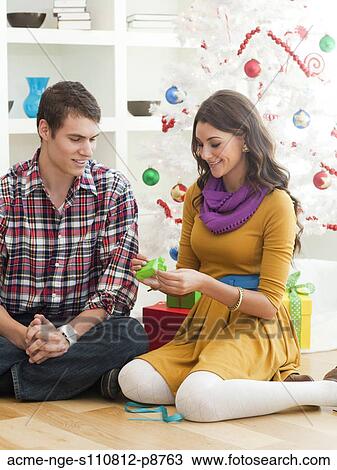 Stock Image - Young couple sitting under Christmas tree, opening Christmas present. Fotosearch