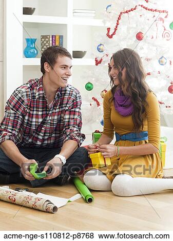Stock Photo - Young couple sitting under Christmas tree, wrapping Christmas presents. Fotosearch