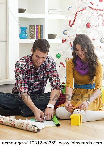 Stock Photo - Young couple sitting under Christmas tree, wrapping Christmas presents. Fotosearch