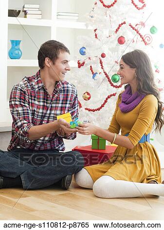 Stock Photography - Young couple sitting under Christmas tree, opening Christmas present. Fotosearch
