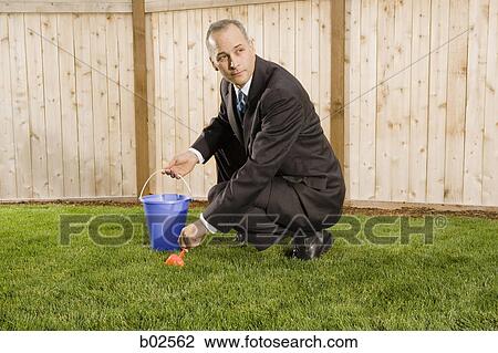 Stock Image - Profile of a businessman crouching on a lawn holding sand pail and shovel. Fotosearch