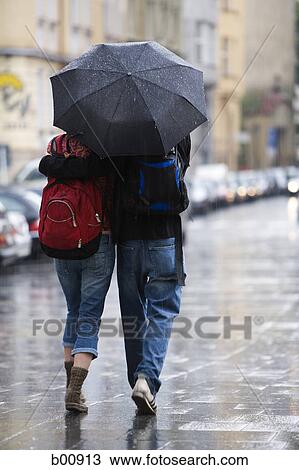 Stock Image - Rear view of a young man walking with a young woman under an umbrella. Fotosearch