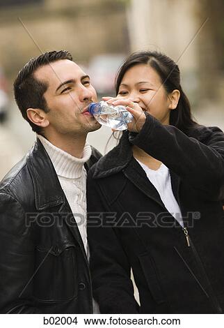 Young man drinking water from a water bottle held by a young woman View Large Photo Image Picture - Young man drinking water from a water bottle held by a young woman. Fotosearch