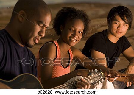 Profile of a young man sitting with two people and playing a guitar View Large Photo Image Stock Image - Profile of a young man sitting with two people and playing a guitar. Fotosearch