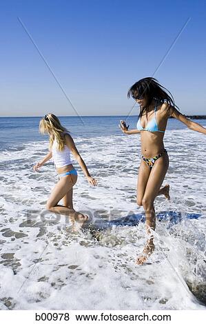 Stock Photo - Profile of two young women running in the water on the beach. Fotosearch