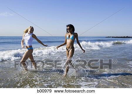 Stock Photo - Two young women running in the water on the beach. Fotosearch