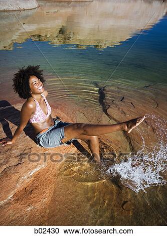 Stock Image - High angle view of a young woman sitting on a rock, splashing water. Fotosearch