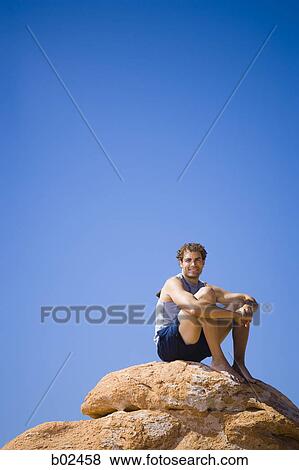 Low angle view of a young man sitting on a rock View Large Photo Image Stock Photo - Low angle view of a young man sitting on a rock. Fotosearch