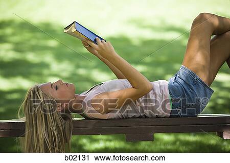 Stock Image - Young woman lying on a park bench and reading a book. Fotosearch