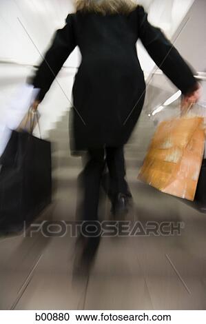 Stock Image - Rear view of a woman running up a staircase with shopping bags. Fotosearch