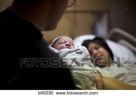Stock Image - Father carrying his newborn baby girl with his wife lying on a bed. Fotosearch