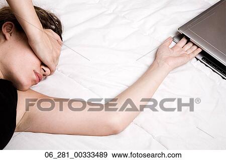 Stock Photo - Above view head and shoulders woman turning her head covering her eyes stretching her arm towards laptop on bed. Fotosearch