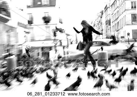 Stock Image - Black and white full length front view woman walking through pigeons on a city street. Fotosearch