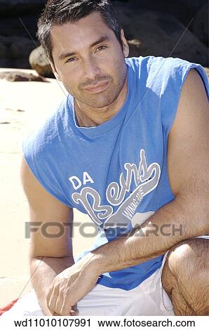 Stock Image - Brunette male looking to camera wearing a logo print cap sleeved shirt and white shorts. Fotosearch