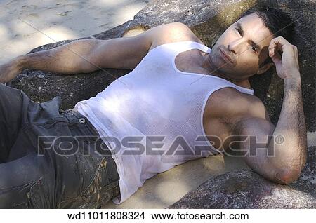 Picture - Brunette male wearing a wet white vest lying back on a rock. Fotosearch