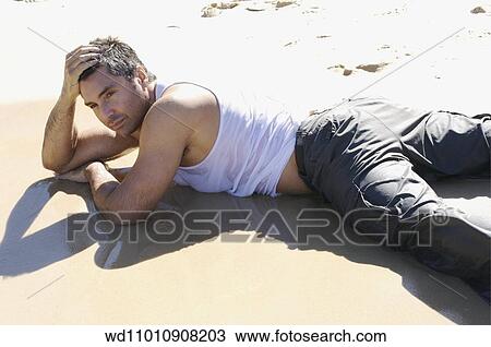 Stock Image - Brunette male wearing a wet white vest and wet grey trousers lying on front on the beach. Fotosearch