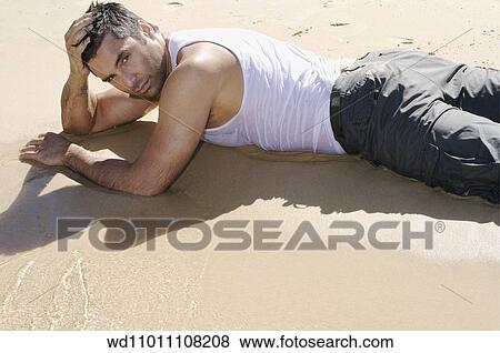 Stock Photo - Brunette male wearing a wet white vest and grey trousers lying on front on the beach. Fotosearch