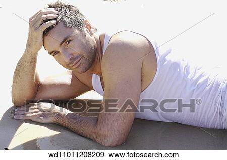 Stock Photo - Brunette male wearing a wet white vest lying on front on the beach. Fotosearch