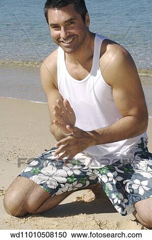 Stock Image - Brunette male wearing a white vest and Hawaiian shorts sitting on knees on the beach. Fotosearch