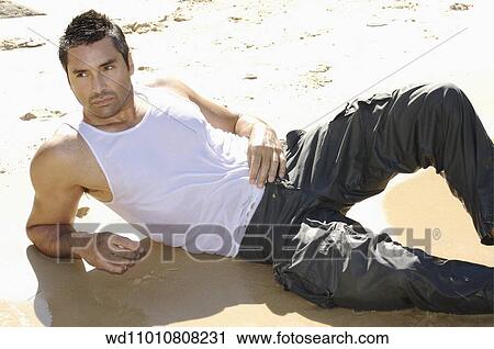 Stock Image - Brunette male wearing a white vest and wet grey trousers lying on the beach. Fotosearch