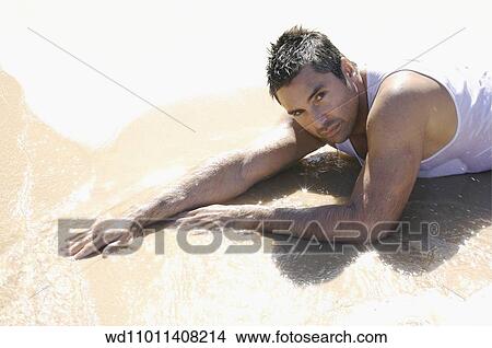 Picture - Brunette male wearing a white vest lying on front in shallow water on the beach. Fotosearch