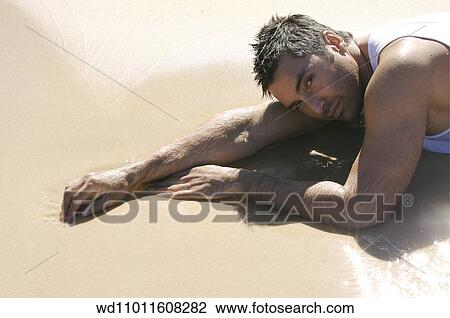 Stock Image - Brunette male wearing a white vest lying on front in shallow water on the beach. Fotosearch