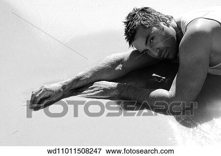 Stock Photo - Brunette male wearing a white vest lying on front in shallow water on the beach. Fotosearch