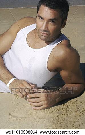 Picture - Brunette male wearing white vest lying on side on the beach. Fotosearch