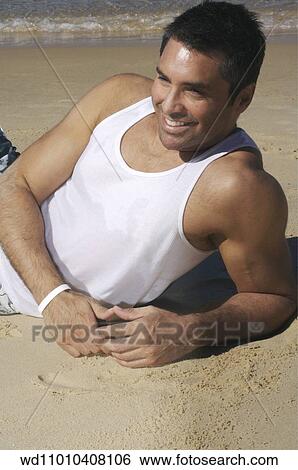 Stock Photograph - Brunette male wearing white vest lying on side on the beach. Fotosearch