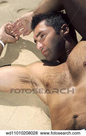 Stock Photo - Brunette male with bare chest and arms above head lying down on the beach. Fotosearch
