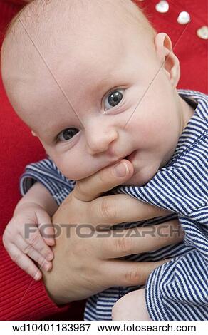 Stock Photo - Close up of a baby being held by someone wearing a red jumper. The baby is wearing a blue and white striped jumper and is looking at the camera.. Fotosearch