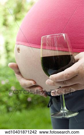Stock Image - Close up of bare stomach of heavily pregnant female holding large glass of red wine in foreground.. Fotosearch