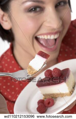 Stock Photograph - Female with straight brunette hair off face wearing red and white spotted shirt out of focus in background holding plate of cheesecake with rasberries and piece on fork looking to camera mouth open smiling showing teeth.. Fotosearch