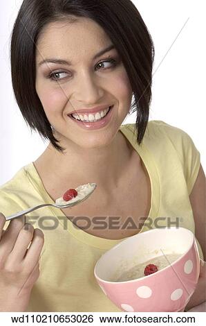 Stock Photograph - Female with straight chin length brunette hair wearing pale yellow t shirt eating porridge with rasberries from pink and white spotted bowl. Fotosearch